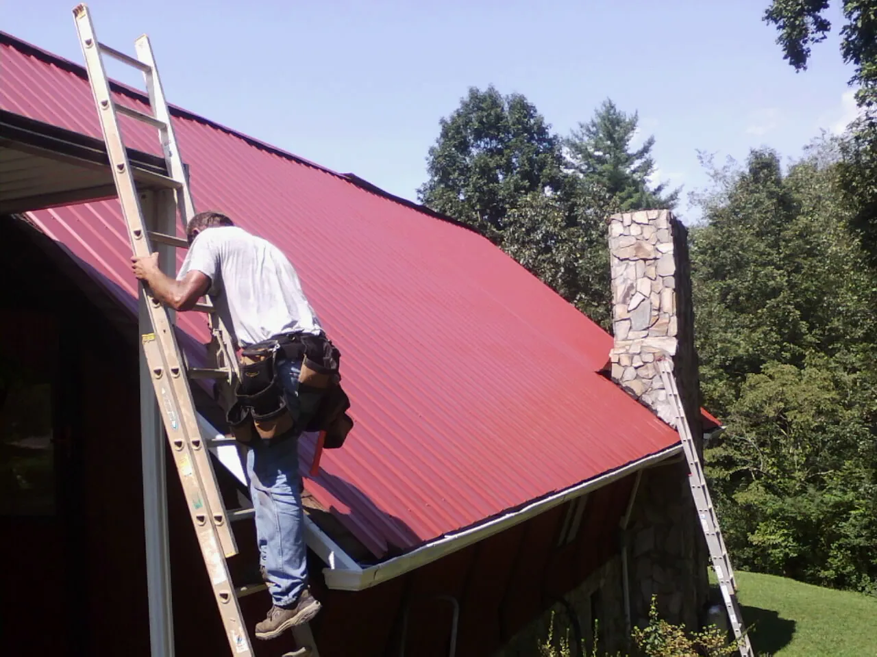 Roofer on a ladder beside a red metal roof and stone chimney — Soffit & Fascia Repair by Roofing Brothers in Belen